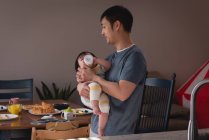 Вид сбоку на счастливого отца-азиата, кормящего маленькую девочку бутылкой с молоком на кухне дома — стоковое фото