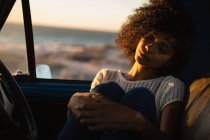 Vista frontal da bela mulher afro-americana pensativa sentada no carro na praia ao pôr do sol — Fotografia de Stock