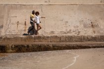 Vue latérale de mignon couple multi ethnique marchant ensemble sur une promenade à la plage par une journée ensoleillée — Photo de stock