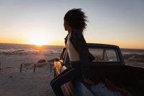 Vista lateral de la hermosa mujer afroamericana sentada en un coche en la playa al atardecer - foto de stock