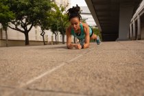 Frontansicht einer jungen Frau mit gemischter Rasse, die Musik über Kopfhörer hört, während sie in der Stadt Plank-Übungen macht — Stockfoto