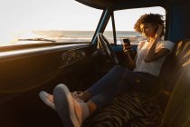 Seitenansicht der schönen jungen afrikanisch-amerikanischen Frau mit Mobiltelefon, während sie sich im Auto am Strand bei Sonnenuntergang lehnt — Stockfoto