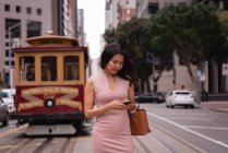 Front view of Asian woman using mobile phone while standing in front of a tram in the street — Stock Photo