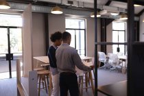 Вид ззаду молодих бізнесменів змішаної раси, які обговорюють синій друк, який тримає чоловік, стоячи в сучасному офісі — стокове фото