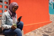 Вид збоку красива змішана раса жінка сміється і використовує мобільний телефон, тримаючи смужку перед будинком на вулиці в сонячний день — стокове фото