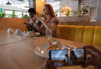 Vista lateral de las amigas de raza mixta tomando fotos de la comida del desayuno con teléfono móvil en el restaurante - foto de stock