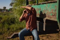 Vista frontal del agricultor caucásico mayor que protege los ojos con el brazo y habla por teléfono móvil mientras está sentado en la parte trasera de su camión en el campo en un día soleado - foto de stock