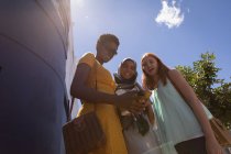 Vista de ángulo bajo de jóvenes amigas de raza mixta usando teléfono móvil en la ciudad en un día soleado - foto de stock