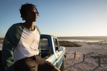 Vista lateral del joven afroamericano sentado en el coche en la playa en un día soleado - foto de stock