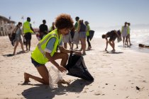 Vista lateral de una joven voluntaria de raza mixta limpiando la playa en un día soleado - foto de stock