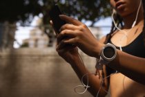 Sección media de la mujer de raza mixta usando el teléfono móvil mientras escucha música en los auriculares de la ciudad - foto de stock