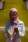 Вид спереди красивой женщины смешанной расы, которая пользуется мобильным телефоном, опираясь на стену в солнечный день — стоковое фото