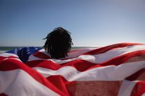Rear view of young African American man holding american flag at beach on a sunny day — Stock Photo