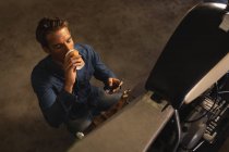 Vista de alto ângulo de caucasiano bicicleta masculina mecânico beber café enquanto usando móvel na garagem — Fotografia de Stock