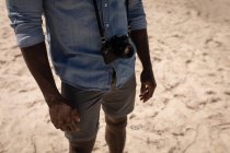 Sección media del hombre de pie con la cámara en la playa en un día soleado - foto de stock