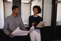Frontansicht glücklicher junger Geschäftsleute mit gemischter Rasse, die im modernen Büro miteinander interagieren, während sie ein digitales Tablet und eine Blaupause in der Hand halten — Stockfoto