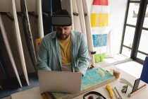 Высокий угол обзора кавказца, работающего на ноутбуке при использовании гарнитуры виртуальной реальности в мастерской — стоковое фото