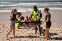 Vue latérale d'un groupe de bénévoles multiethniques nettoyant la plage à l'aide d'une passoire spéciale tandis que les autres bénévoles sont assis en position accroupie derrière eux par une journée ensoleillée — Photo de stock