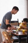 Вид сбоку отца-азиата, подающего завтрак своим детям за обеденным столом на кухне дома — стоковое фото
