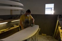 Frontansicht eines kaukasischen Mannes, der in einer Werkstatt Surfbrett mit Maschine formt und dabei eine Schutzmaske trägt — Stockfoto