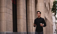 Front view of handsome Asian man standing on street while holding mobile phone and coffee in hand — Stock Photo