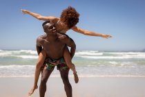 Vista frontal do homem afro-americano feliz e mulher de raça mista em pé na posição de piggyback na praia em um dia ensolarado — Fotografia de Stock