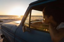 Вид сбоку красивой молодой афроамериканки, мечтающей, сидя в машине на пляже на закате — стоковое фото