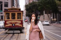 Vue de face de femme asiatique réfléchie debout devant un tramway dans la rue tout en buvant du café — Photo de stock