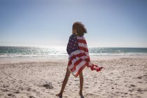 Rückansicht einer jungen Frau, die an einem sonnigen Tag am Strand steht und eine amerikanische Flagge trägt — Stockfoto
