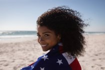Porträt einer jungen afrikanisch-amerikanischen Frau mit amerikanischer Flagge an einem sonnigen Tag am Strand — Stockfoto