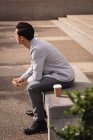 Vue latérale du jeune homme asiatique prenant son café et son petit déjeuner assis sur un banc en pierre — Photo de stock