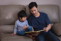 Vista frontal do pai asiático lendo livro de história para seu filho enquanto sentado no sofá em casa — Fotografia de Stock