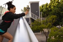 Vista lateral de la joven mujer de raza mixta utilizando el teléfono móvil en el puente de la ciudad - foto de stock