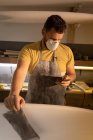 Front view of Caucasian man with mouth protection mask using mobile phone while cleaning surfboard in a workshop. — Stock Photo