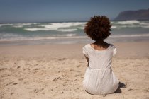 Вид спереду змішаної раси жінка в білій сукні, сидячи на піску, розслабляючись на пляжі на сонячному — стокове фото