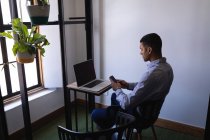Vista lateral del joven hombre de negocios de raza mixta que utiliza el teléfono móvil mientras trabaja en el ordenador portátil en el escritorio en la oficina - foto de stock