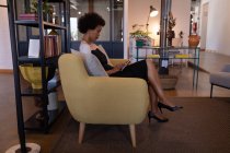 Вид сбоку красивой бизнесвумен смешанной расы, использующей цифровой планшет в современном офисе, пока она сидит на современном сиденье на фоне мебели на заднем плане — стоковое фото