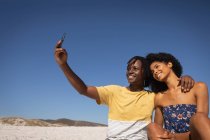 Vue de face de heureux jeune couple afro-américain prenant selfie tout en étant assis à la plage par une journée ensoleillée — Photo de stock
