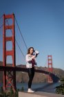 Vista laterale di bella donna che prende selfie vicino al ponte sospeso — Foto stock