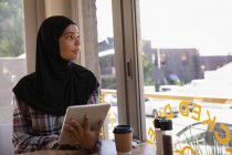 Front view of thoughtful young woman in hijab using digital tablet in a cafe — Stock Photo