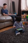 Vista frontale del padre asiatico che usa il suo cellulare sul divano mentre suo figlio usa un tablet digitale a casa — Foto stock