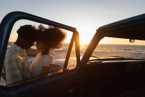 Vue latérale du couple romantique afro-américain debout à la plage près de la voiture au coucher du soleil — Photo de stock
