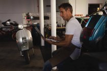 Vue latérale du mécanicien de vélo caucasien en utilisant un ordinateur portable tout en étant assis dans le garage — Photo de stock