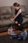 Vue de face d'une mère asiatique utilisant un ordinateur portable et assise sur le canapé pendant que les enfants jouent sur un tapis avec une tablette numérique à la maison — Photo de stock