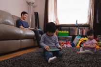 Low angle view of Asian father using his mobile phone on the sofa while his son is using a digital tablet and daughter playing at home — Stock Photo