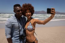 Вид спереди симпатичной многоэтнической пары, делающей селфи с мобильным телефоном на пляже — стоковое фото