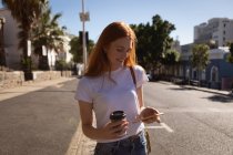 Vista frontal de la hermosa joven mujer caucásica usando el teléfono móvil mientras toma café en la calle en un día soleado - foto de stock