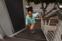 Hochwinkelaufnahme einer jungen Frau mit gemischter Rasse, die Musik über Kopfhörer hört, während sie auf Treppen in der Stadt trainiert — Stockfoto