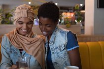 Vista frontal de belas amigas afro-americanas usando telefone celular enquanto estão sentadas no restaurante — Fotografia de Stock
