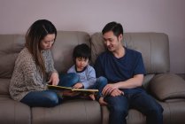 Vue de face du livre de lecture de famille asiatique heureux tout en étant assis ensemble sur le canapé à la maison — Photo de stock